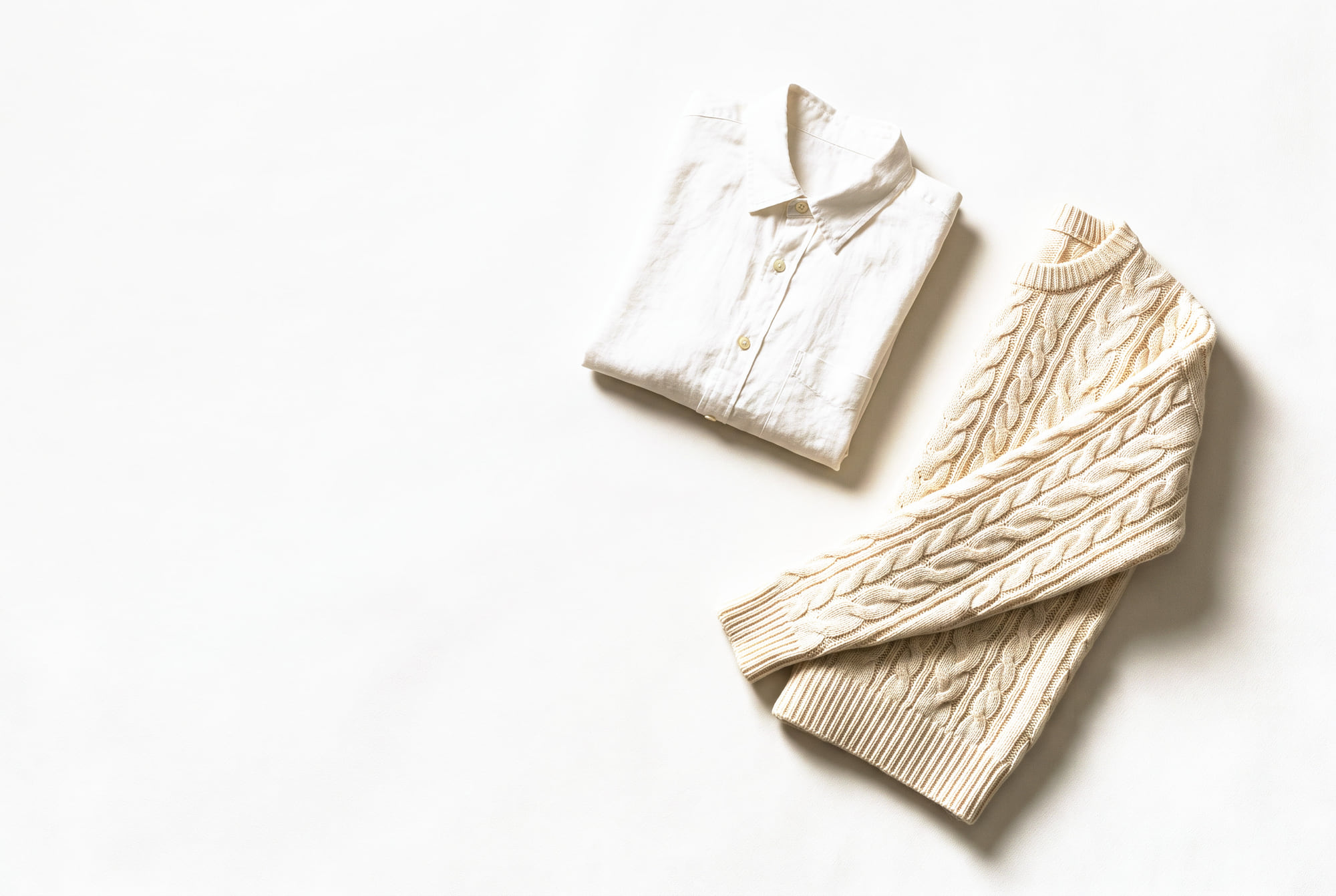 A folded white linen shirt beside an ivory cable-knit jumper with one sleeve laid out, on a pale ground.